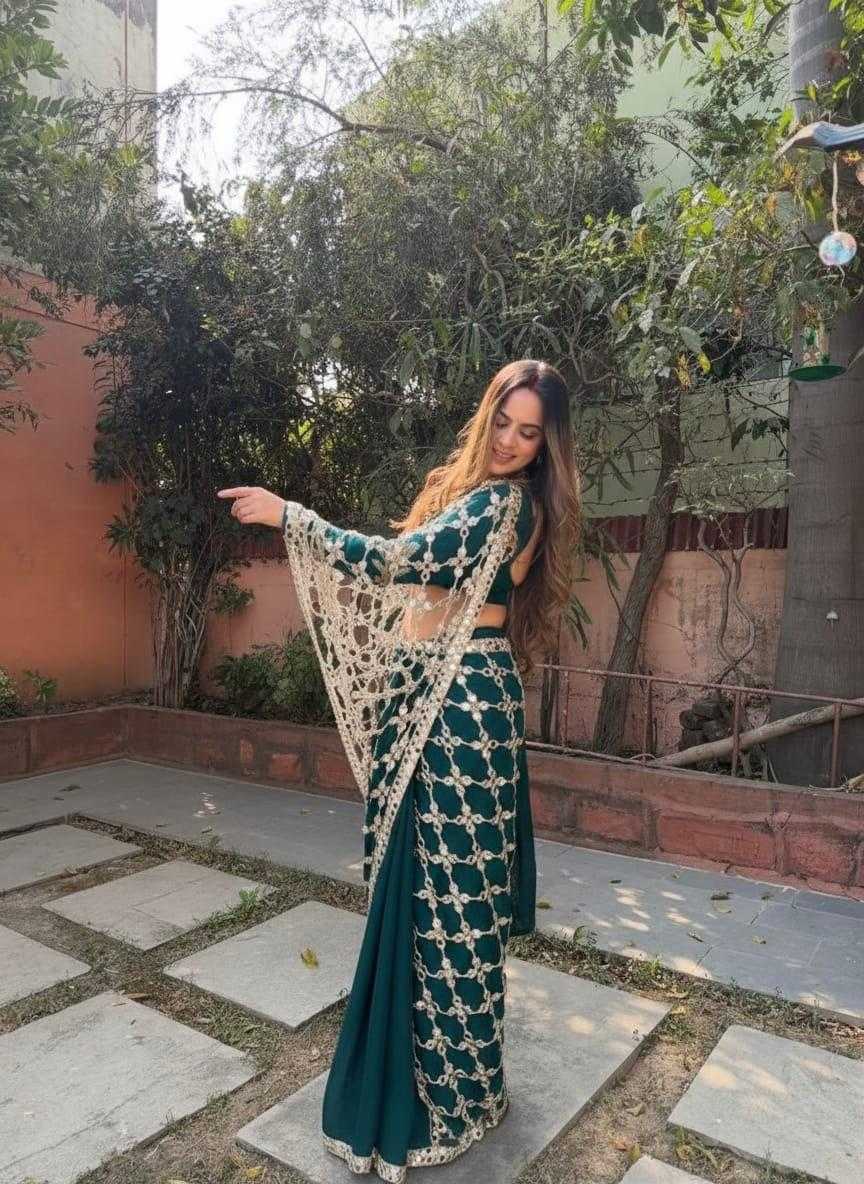 A woman wearing a dark teal green and gold embroidered traditional Indian outfit with a matching dupatta, standing outdoors on a stone patio with trees and a wall in the background.