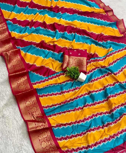 A striped sari laid flat on a white textured surface, featuring alternating bands of turquoise blue, mustard yellow, and deep red with a wide red and gold zari border. A matching red blouse piece with gold detailing is folded and placed in the center, alongside a small green plant sprig.