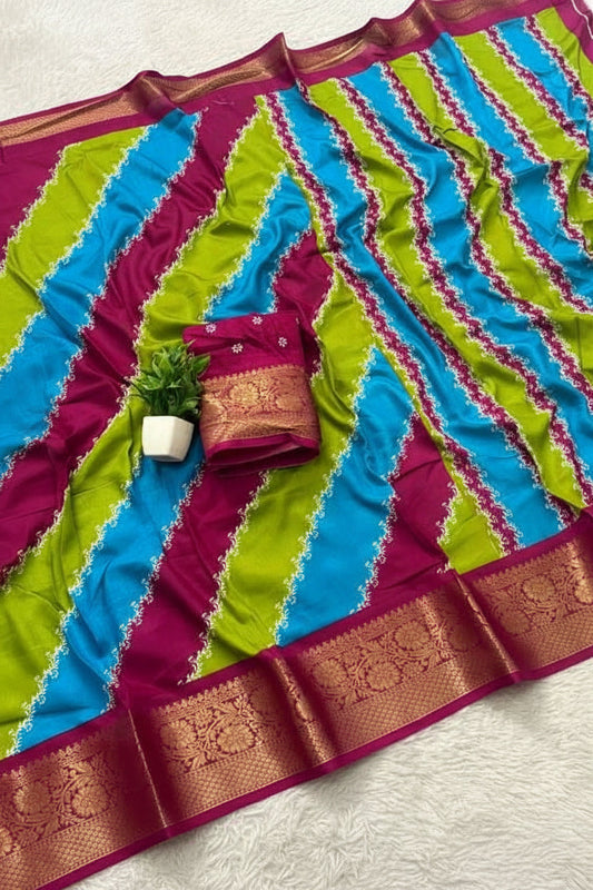 A colorful striped fabric, likely a saree or scarf, featuring diagonal bands of turquoise blue, lime green, and magenta pink with ornate gold zari borders. A matching folded blouse piece with a gold border is placed on top next to a small potted plant.