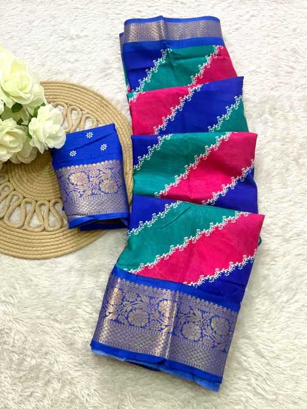 A folded multicolor saree or fabric set displayed on a white textured surface, featuring blue, green, and pink sections with ornate silver zari borders and floral motifs. A matching blue fabric piece with a silver border is placed beside it on a woven mat with white flowers.