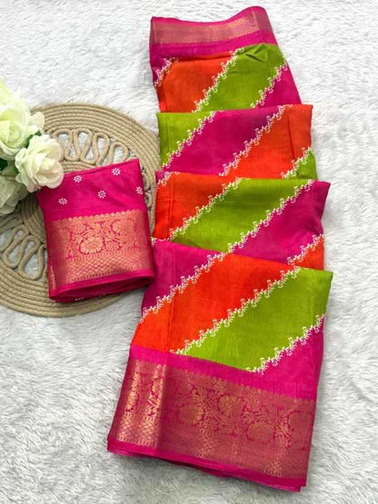 A folded and draped saree with vibrant diagonal stripes of pink, orange, and lime green, featuring a wide pink border with gold zari embroidery, displayed on a white textured background next to a woven mat and white flowers.