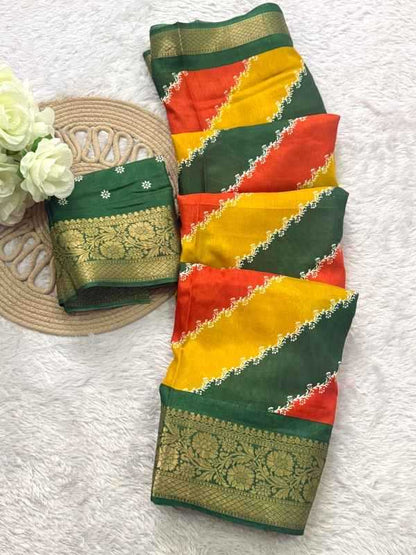 A folded multicolor saree or fabric piece displayed on a white textured surface, with a matching folded section placed beside it on a round woven mat and white flowers. The fabric features diagonal bands of dark green, bright yellow, and orange-red, with ornate gold zari borders and white embroidered details.