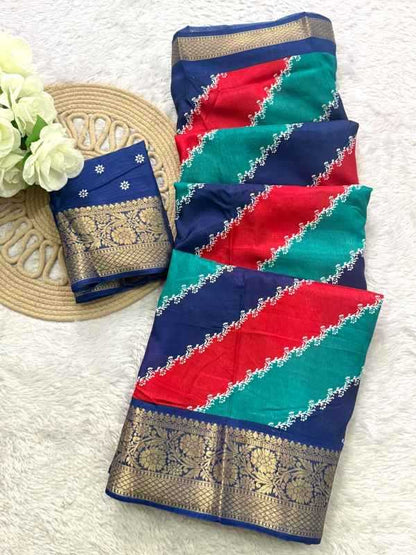 A folded multicolor saree with navy blue, red, and teal diagonal stripes, decorated with intricate gold zari borders and white floral embroidery, displayed on a white textured background next to a woven mat and white flowers.