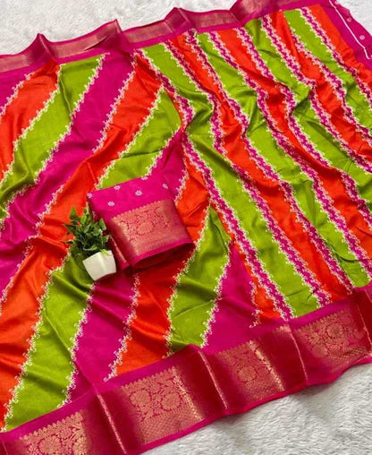 A vibrant striped saree with diagonal bands of lime green, orange, and magenta pink, featuring a wide gold zari border and a matching pink blouse piece with a gold border, styled with a small potted plant on a white textured background.