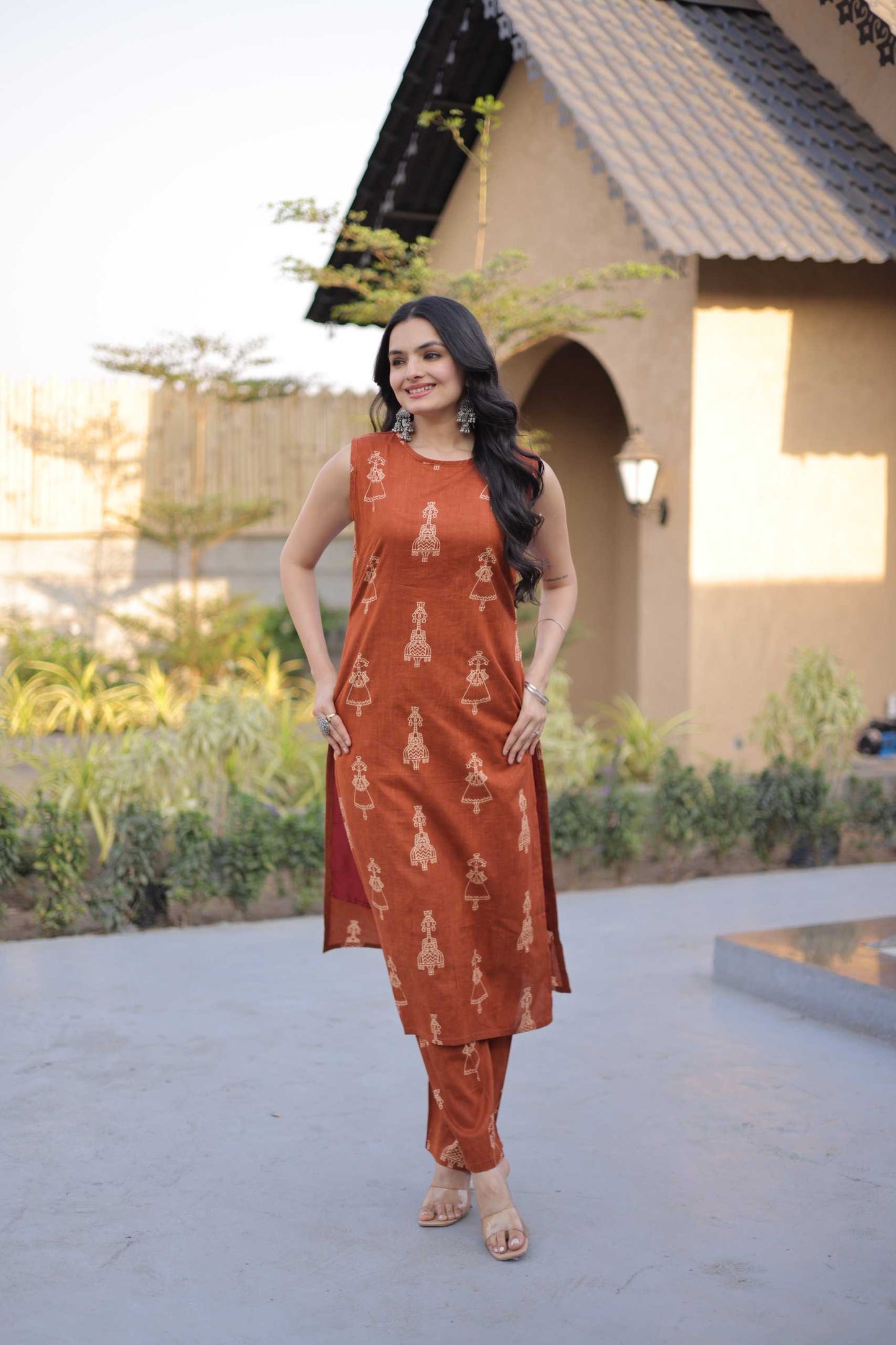 Woman in a rust-colored traditional outfit standing outdoors with a building and greenery in the background.