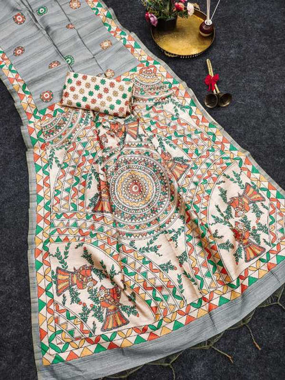 A printed cotton saree or fabric set laid flat on a dark textured surface, featuring a light gray base with a large cream panel decorated with colorful mandala and floral motifs in green, orange, red, and yellow. A matching patterned fabric piece and a small decorative tray with flowers are placed nearby.