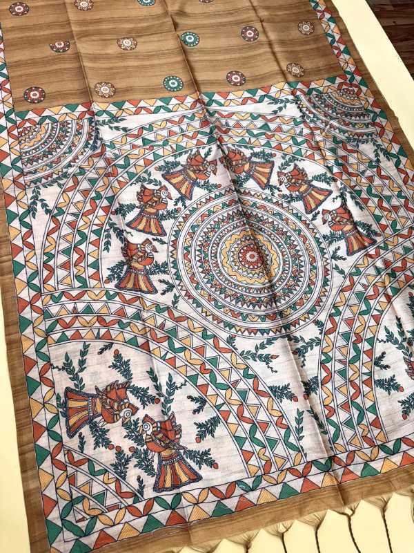 A folded textile product, likely a sari or scarf, featuring a traditional Indian hand-painted style design. The fabric has a warm brown base with a large white central panel decorated with intricate mandalas, floral motifs, and stylized human figures in orange, green, and blue, finished with a border of geometric patterns and tassels.