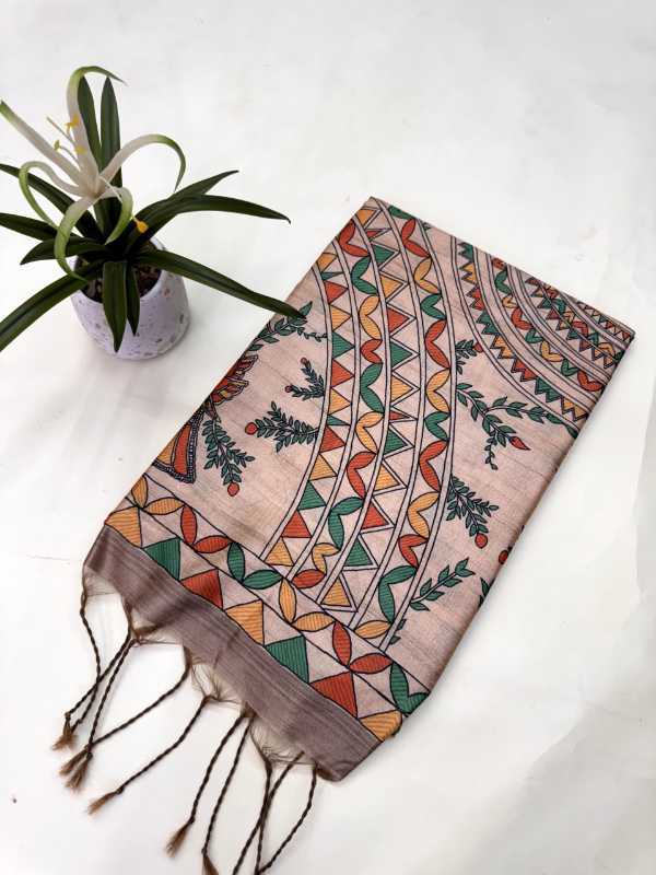 A rectangular scarf with fringed tassels, featuring a traditional Indian Kalamkari style print in earthy tones of orange, green, brown, and beige on a light pinkish-beige base. The scarf is displayed diagonally on a white marble surface next to a small potted plant.
