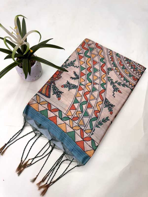 A folded patterned scarf with tassels, displayed on a white fabric background next to a small potted plant. The scarf features a light pink base with geometric tribal patterns in orange, green, and blue, finished with a solid blue border and black tassels.