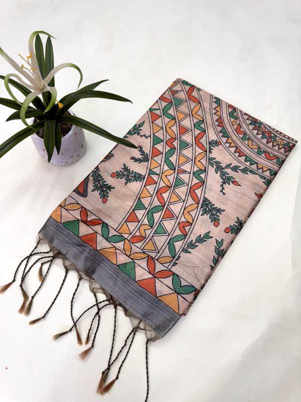 A rectangular scarf with a multicolor geometric and floral mandala pattern on a light pink base, featuring black tassels on one end, displayed on a white surface next to a small potted plant.
