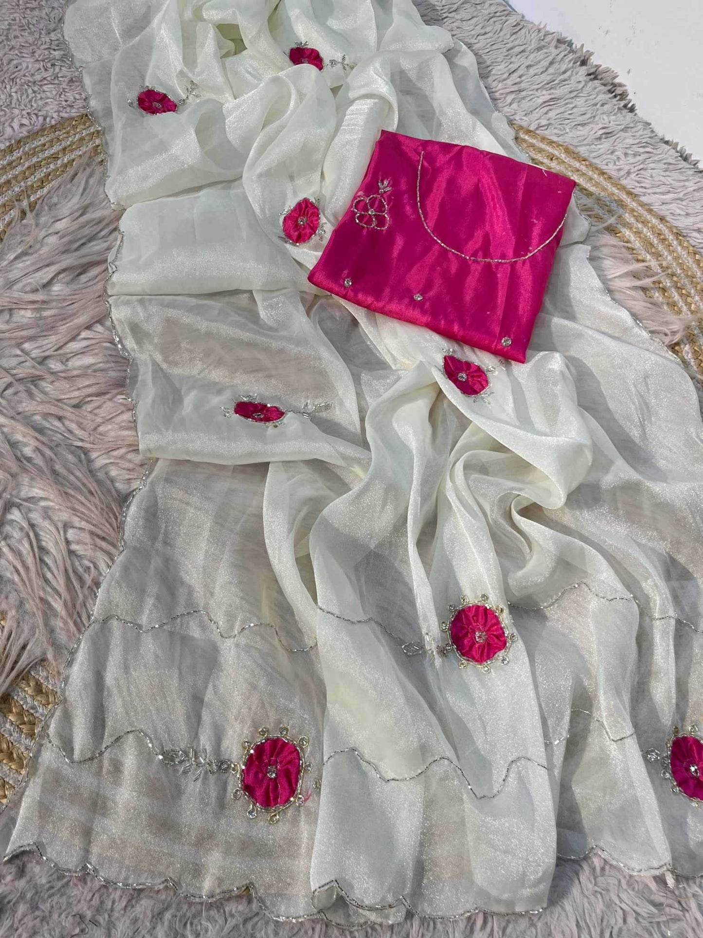An off-white semi-sheer organza saree with vibrant magenta pink floral embroidery and silver mirror work borders, displayed with a matching bright pink blouse piece on a textured rug.