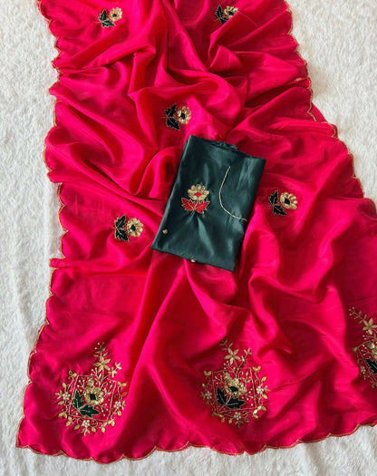 A bright pink silk saree or dupatta with gold and multicolor floral embroidery, laid out on a white textured fabric background with a dark green fabric piece placed in the center.