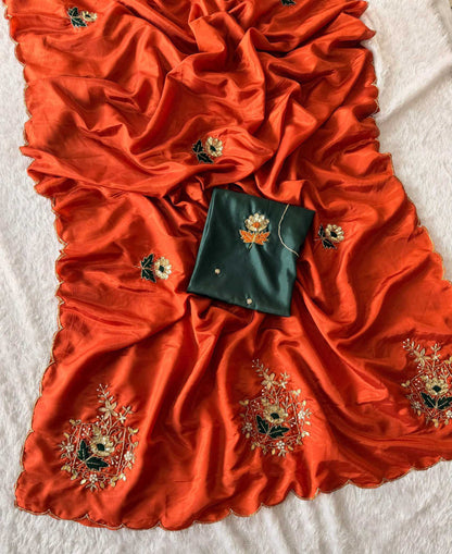 A bright orange-red silk saree or dupatta with gold and green floral embroidery, laid out on a white textured fabric with a dark green fabric piece placed in the center.
