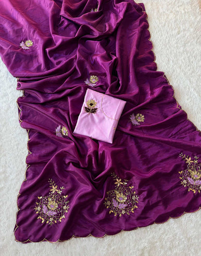 A draped magenta purple silk saree or fabric with gold and green floral embroidery, featuring a matching folded light pink blouse piece with embroidery, laid out on a white textured background.