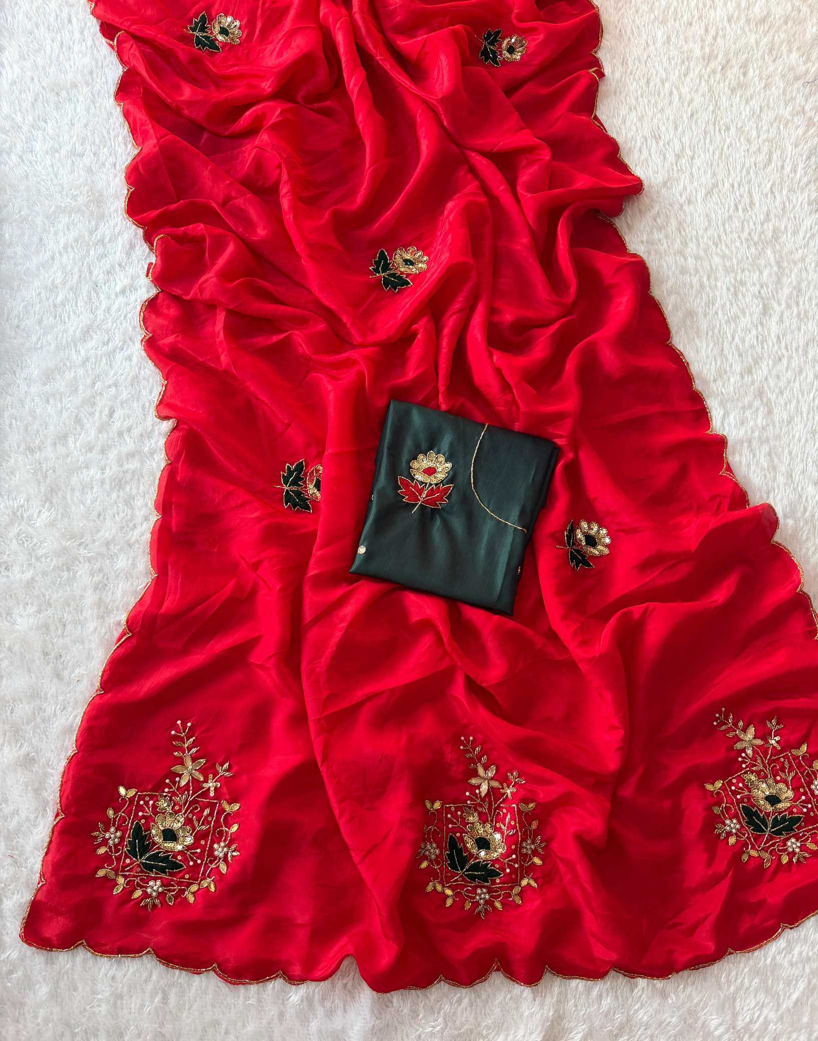 A bright red draped fabric, likely a dupatta or sari, decorated with gold and black floral embroidery, featuring a matching black fabric pouch with a floral design, laid out on a white textured background.