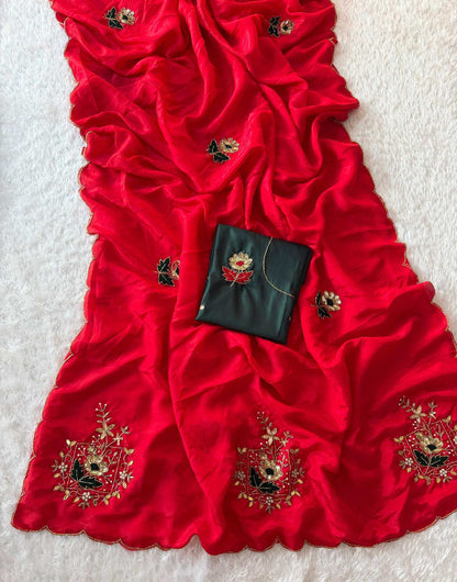A bright red draped fabric, likely a dupatta or sari, decorated with gold and black floral embroidery, featuring a matching black fabric pouch with a floral design, laid out on a white textured background.