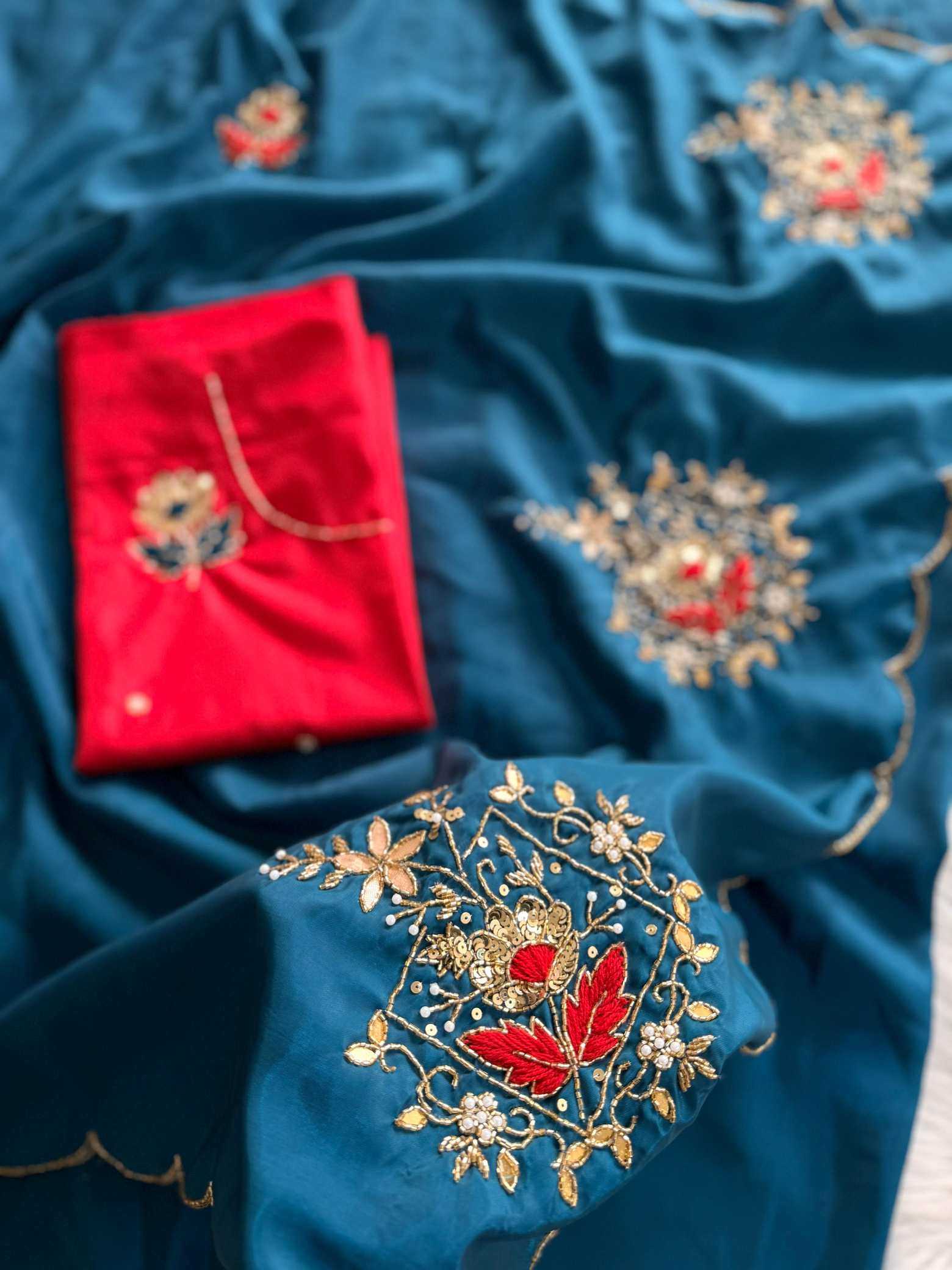 A close-up of a teal blue embroidered fabric, likely a saree or textile, featuring detailed gold floral embroidery with red and white accents. A folded bright red fabric piece with gold embroidery is placed on top of the teal cloth.