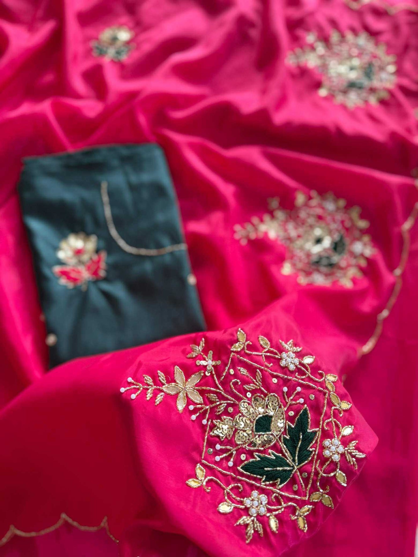 A close-up of a bright pink fabric garment or textile with ornate gold floral embroidery and a small dark teal fabric piece with embroidered detail, arranged on a matching pink satin background.