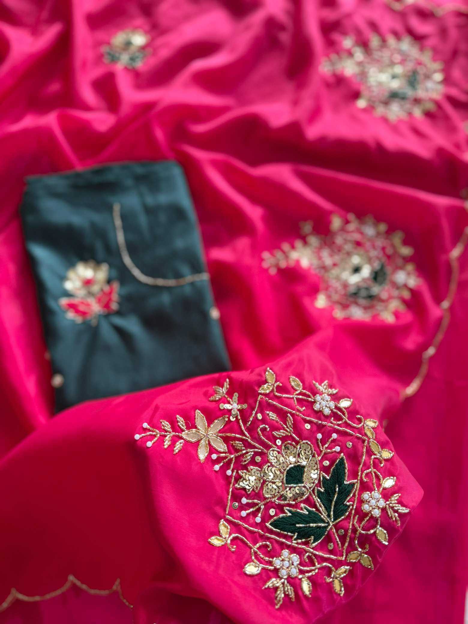 A close-up of a bright pink fabric garment or textile with ornate gold floral embroidery and a small dark teal fabric piece with embroidered detail, arranged on a matching pink satin background.