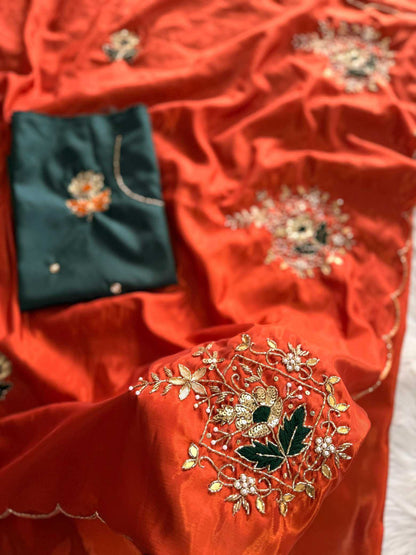 A close-up of a richly textured orange-red fabric, likely a traditional saree or dupatta, featuring intricate gold floral embroidery with green and white accents. A folded dark teal-green fabric piece is tucked into the folds on the left, showcasing a matching embroidered motif.
