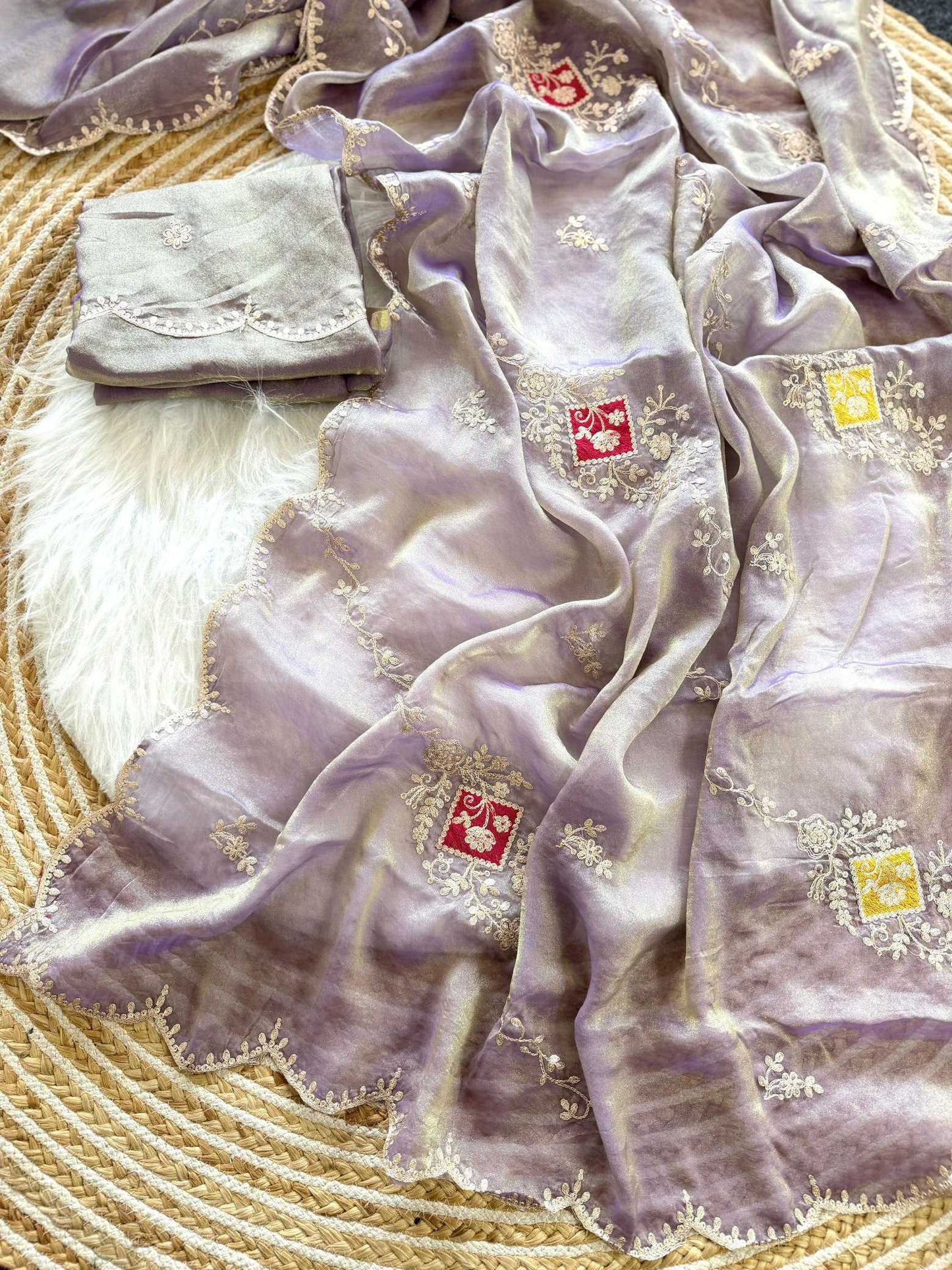 A lavender purple embroidered scarf with scalloped edges and small red and yellow embroidered squares, draped over a white faux fur rug on a woven straw mat.