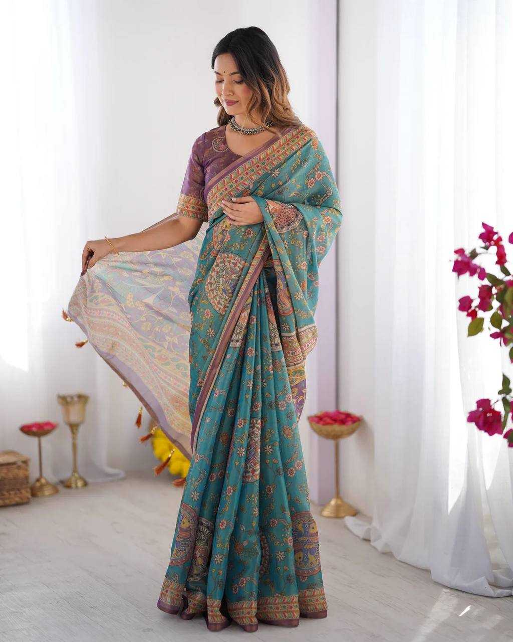 Woman wearing a teal saree with a pattern, standing indoors with white curtains and decorative items in the background.