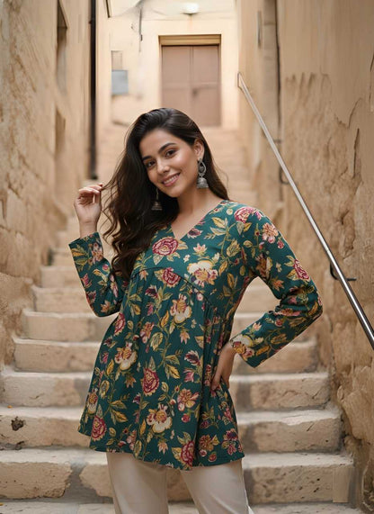 A woman posing on stone stairs wearing a long-sleeve V-neck tunic top with a green floral print and cream pants.