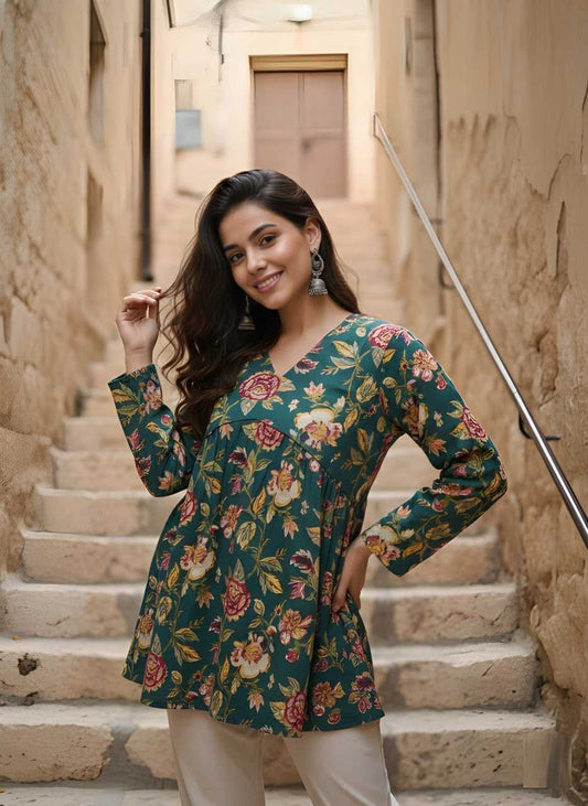 A woman posing on stone stairs wearing a long-sleeve V-neck tunic top with a green floral print and cream pants.