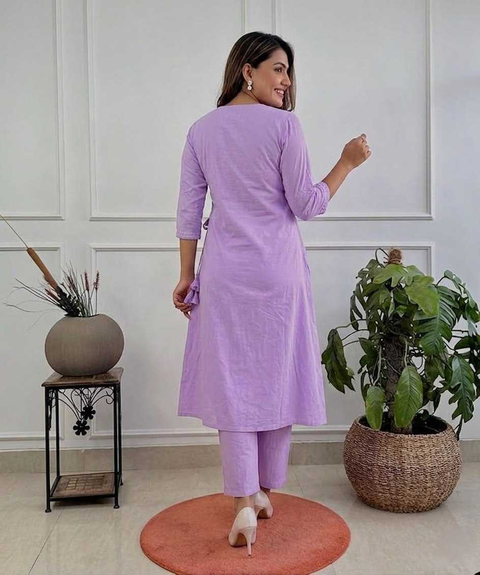 Woman in a lavender outfit standing in a room with plants and a white wall.