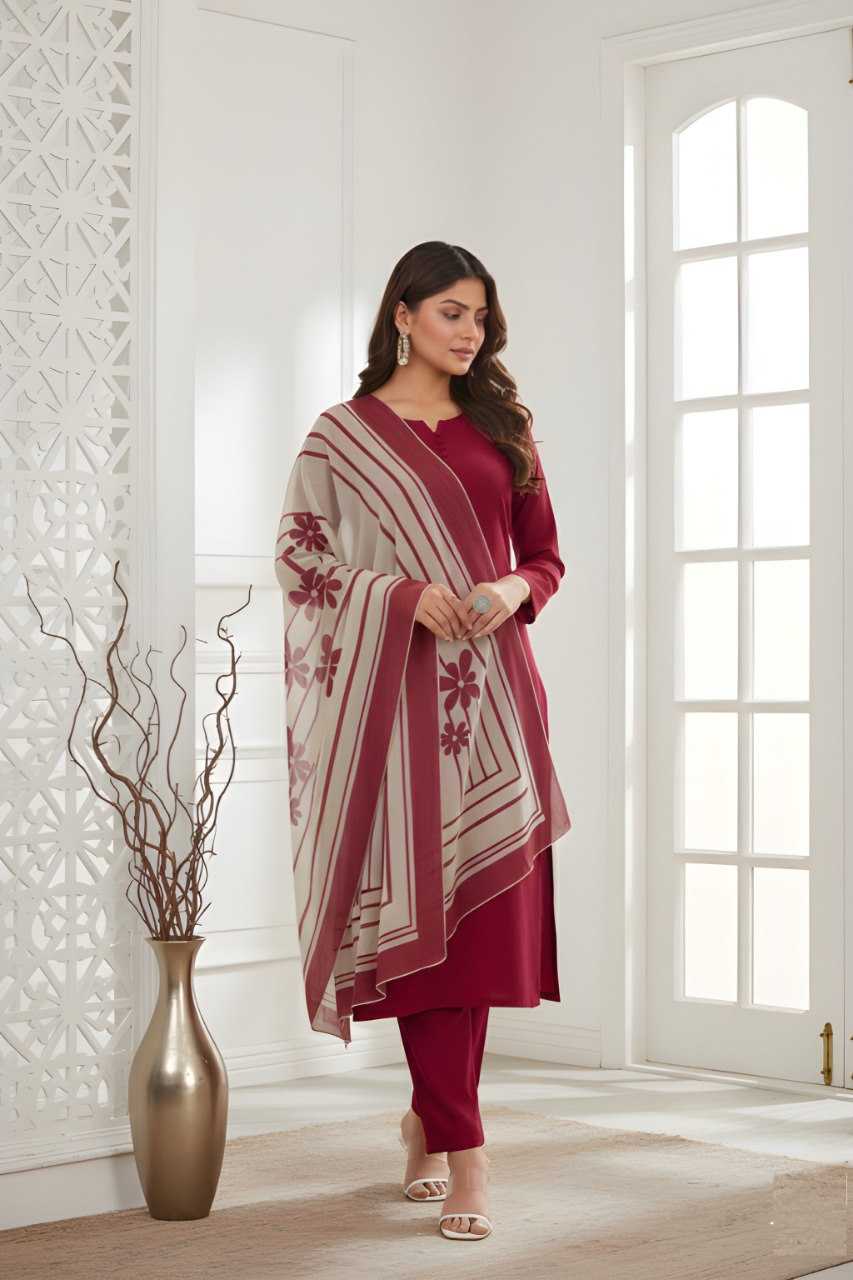 A woman models a three-piece maroon kurta set with matching trousers and a beige and maroon striped shawl featuring red floral motifs, standing in a bright, modern room with white walls and a decorative gold vase.