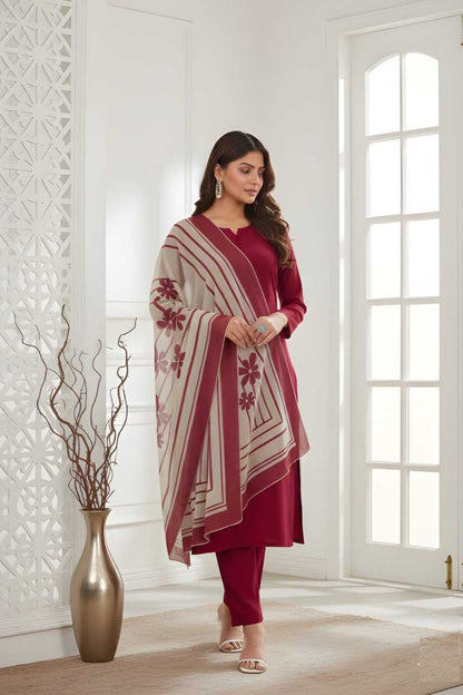 A woman models a three-piece maroon kurta set with matching trousers and a beige and maroon striped shawl featuring red floral motifs, standing in a bright, modern room with white walls and a decorative gold vase.