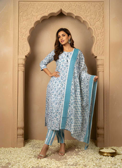 A woman models a traditional Indian salwar kameez suit set in front of an ornate beige archway. The set features a light blue and white floral print kurta with a solid light blue border, matching printed trousers, and a coordinating dupatta.