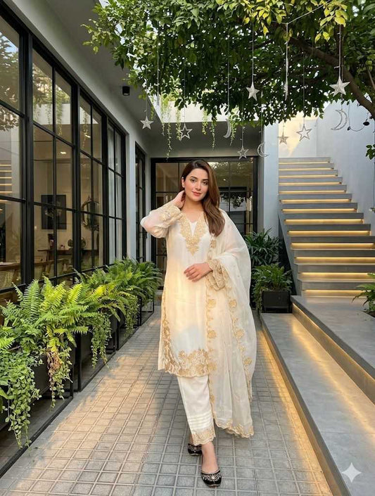 Woman in a white and gold traditional outfit standing in a modern indoor setting with plants and stairs.