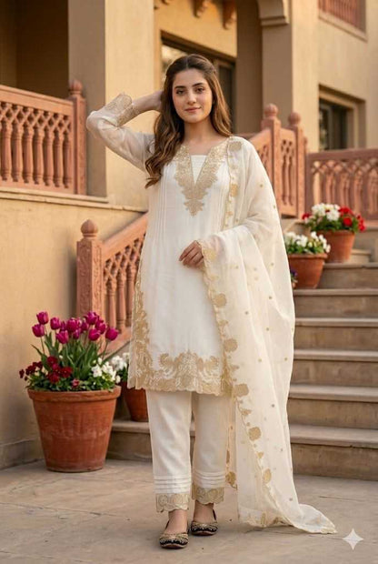 Woman in a white and gold traditional outfit standing outdoors with decorative plants and a building in the background.