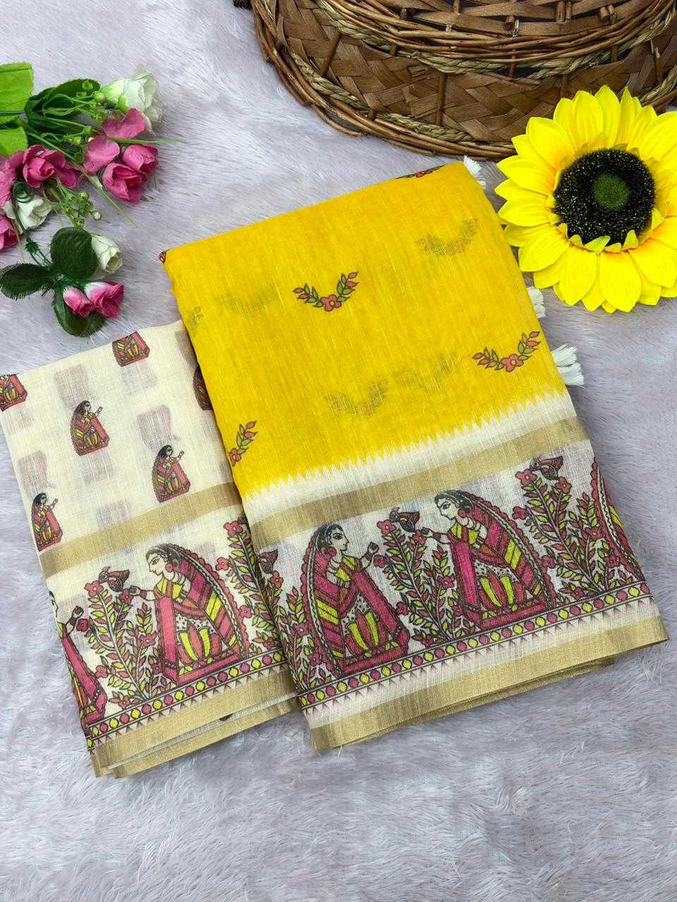 A folded yellow and beige cotton saree with traditional hand-block printed motifs of seated women and floral patterns, styled on a gray faux fur surface with decorative flowers and a basket.