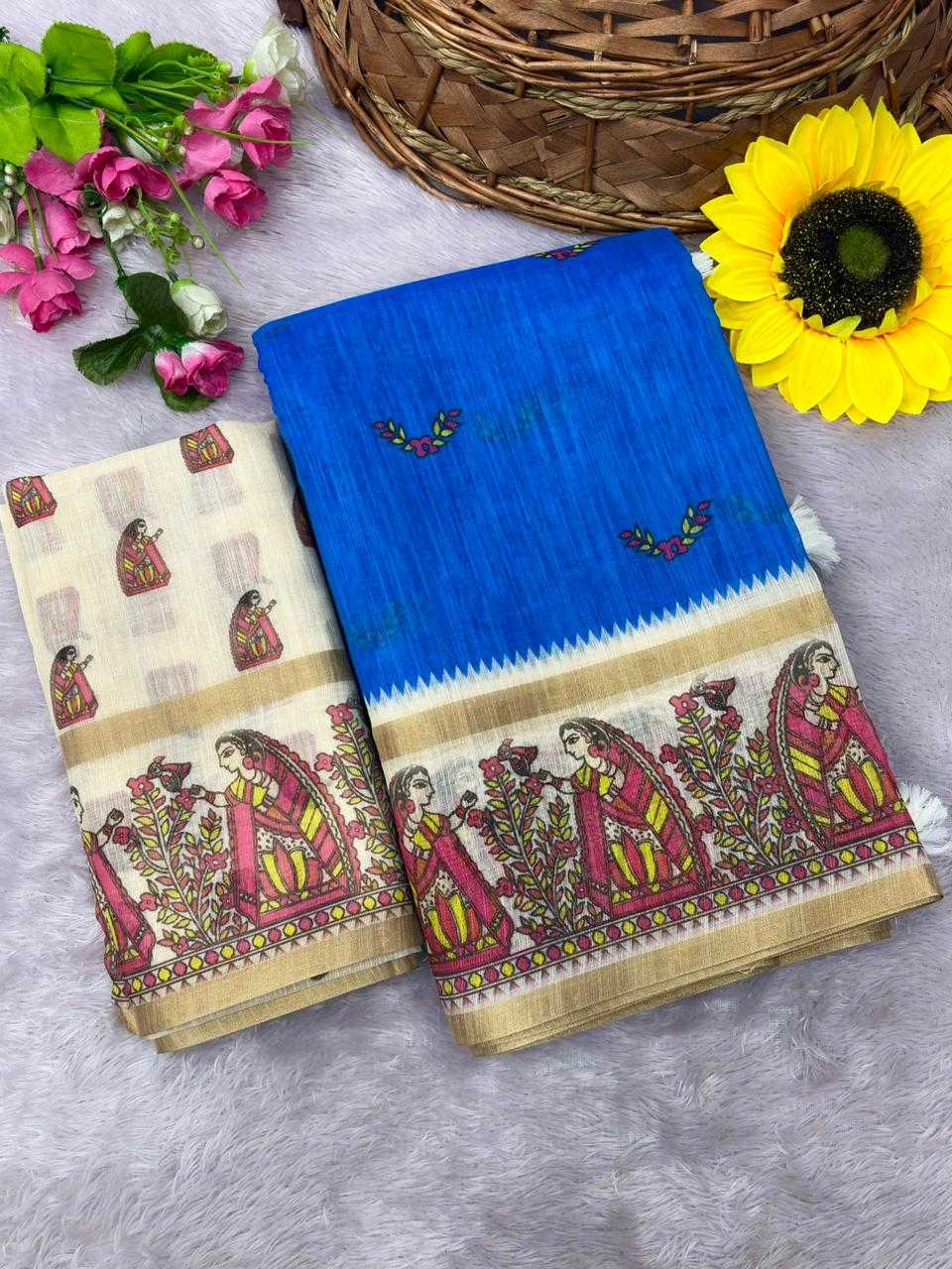 A folded cotton saree displayed on a gray faux fur surface, decorated with pink flowers, a yellow sunflower, and a wicker basket. The saree features a vibrant blue upper section with a zigzag border and a beige lower section with a detailed multicolor traditional illustration.