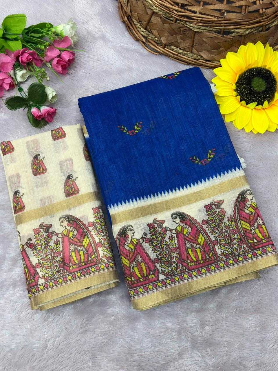 Two folded pieces of printed fabric laid on a light gray textured surface, with decorative flowers and a woven basket around them. One piece is bright royal blue with a wide beige border featuring multicolored traditional-style figures and floral motifs; the other is a light beige/cream piece with similar printed figures and a blue border.