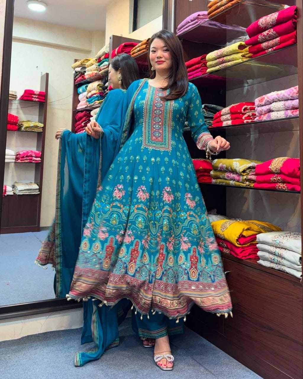 Woman in a blue embroidered traditional outfit standing in a store with folded fabrics in the background.