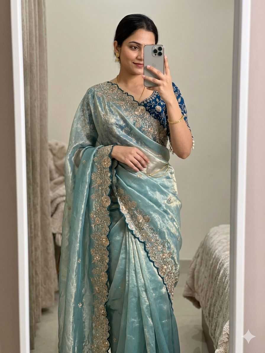 Woman wearing a light blue saree with intricate designs, taking a mirror selfie.