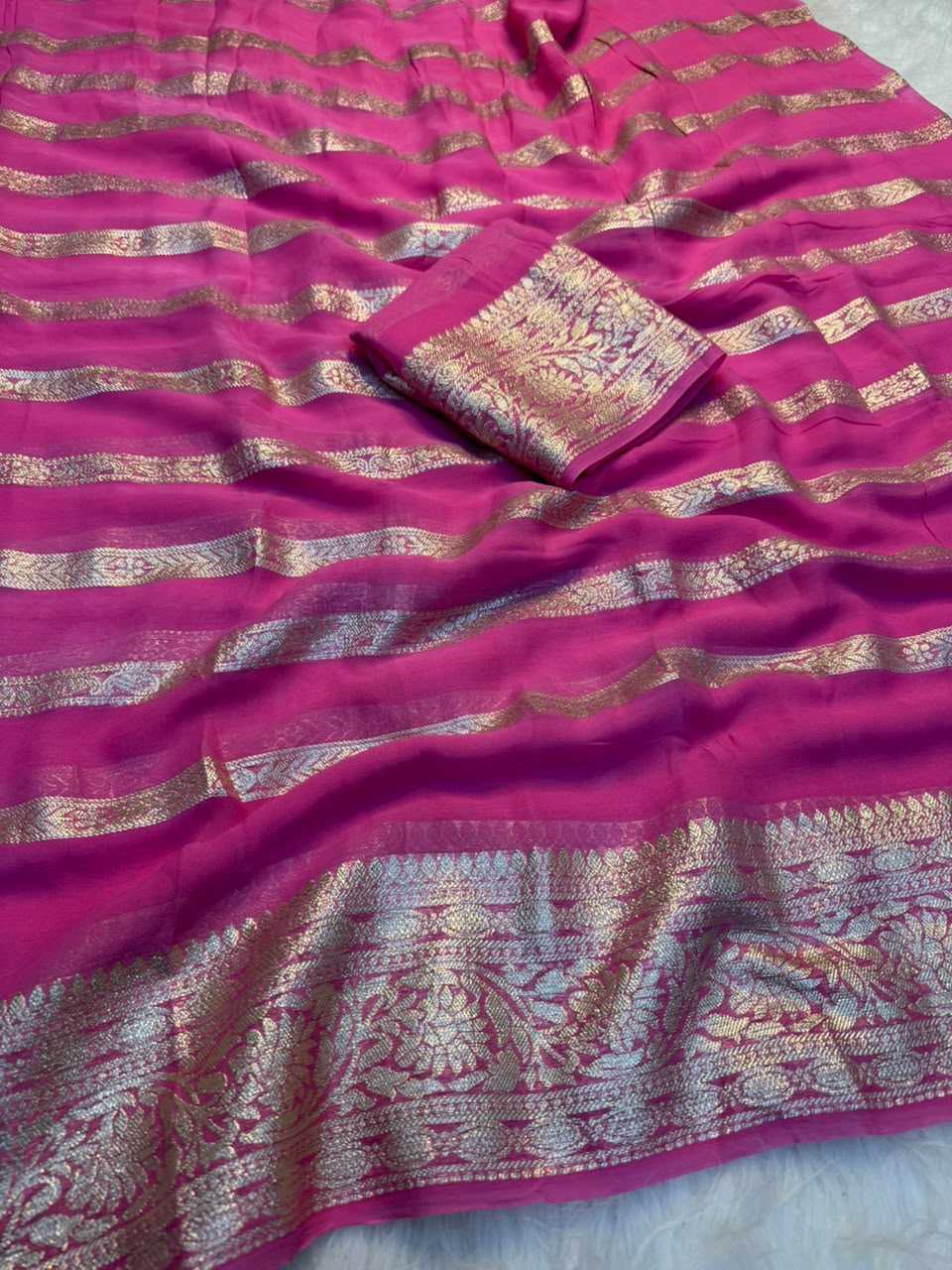 A close-up of a magenta pink fabric, likely a saree or dupatta, featuring horizontal metallic gold and silver zari woven stripes and a detailed border.