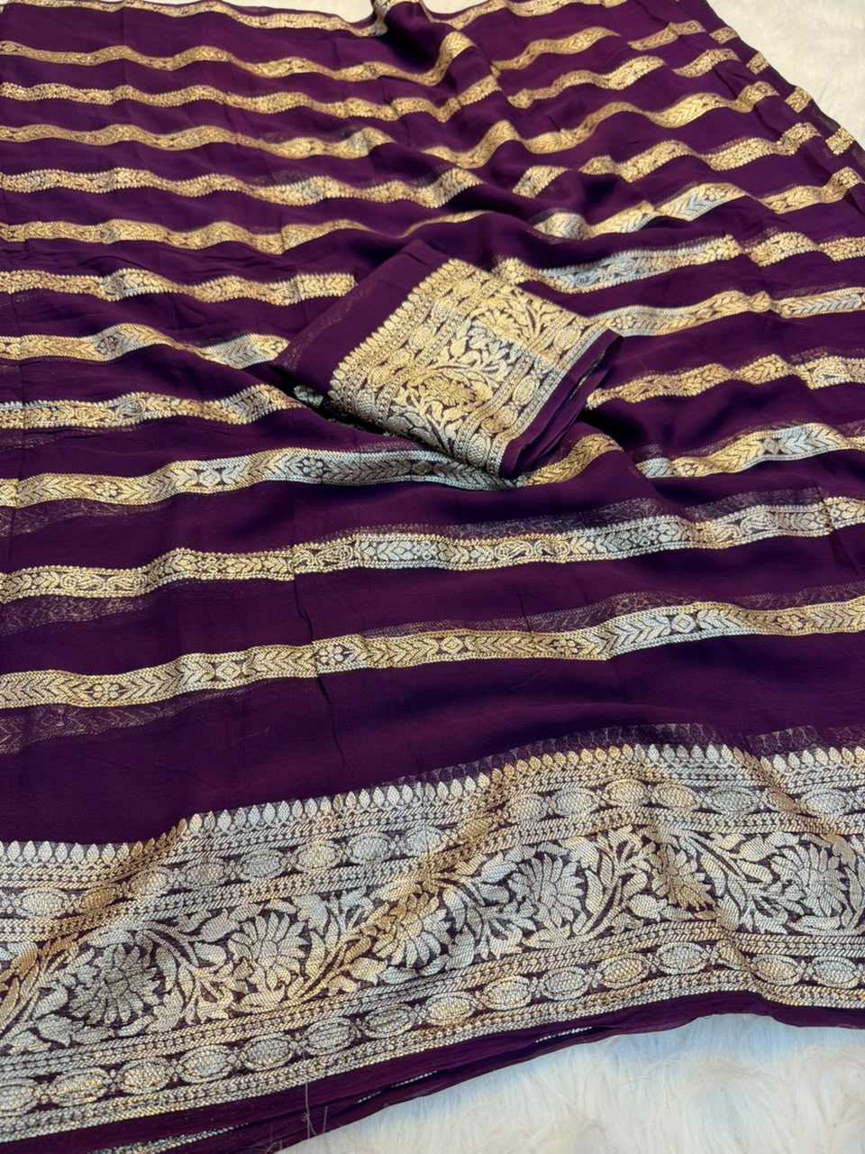 A close-up of a purple fabric, likely a saree or scarf, featuring horizontal metallic gold and silver zari stripes and a detailed floral border.