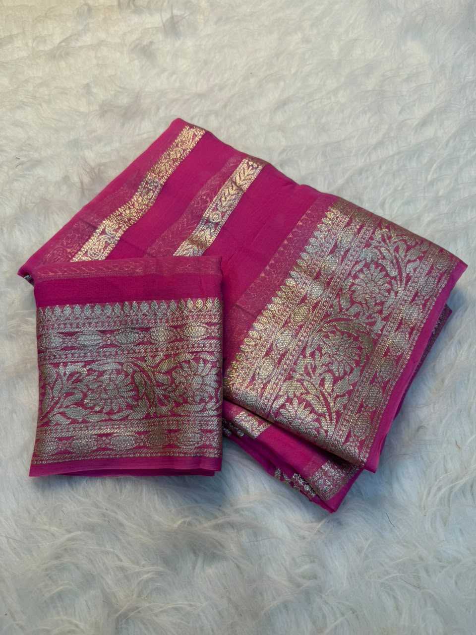 A folded magenta pink silk saree with intricate gold zari brocade patterns, displayed on a white faux fur surface.