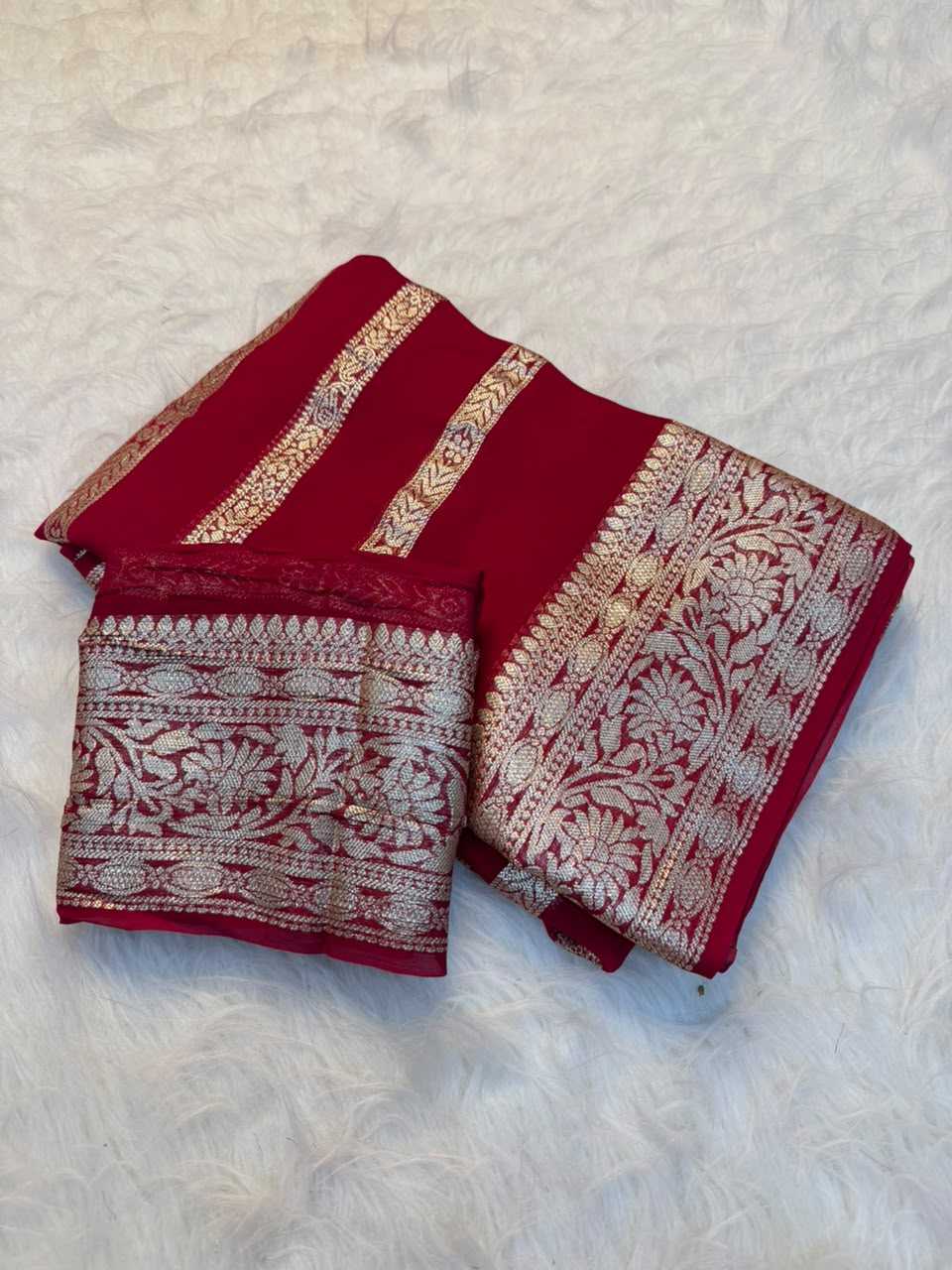A folded deep red saree or fabric piece with ornate silver and gold woven floral borders, displayed on a white faux fur surface.