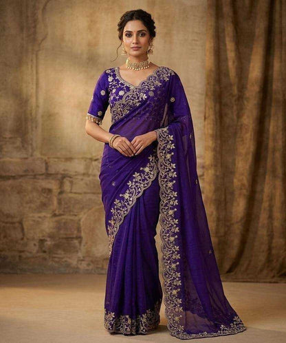 Woman wearing a purple saree with silver embroidery against a textured brown background