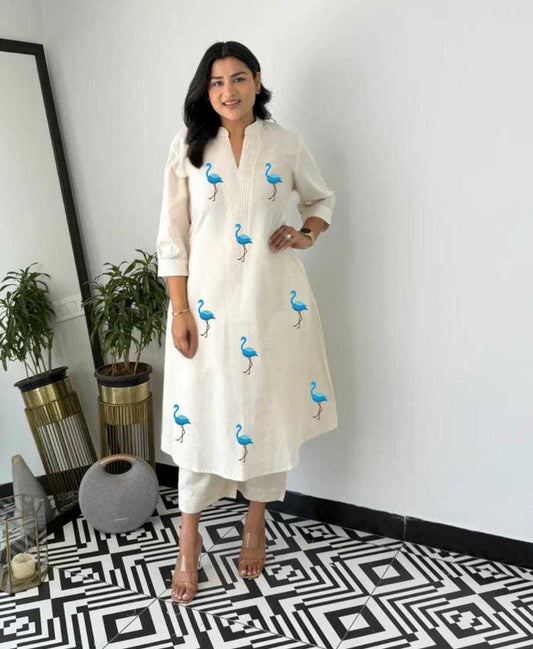 Woman wearing a white kurta with blue bird patterns in a room with decorative plants and a mirror.