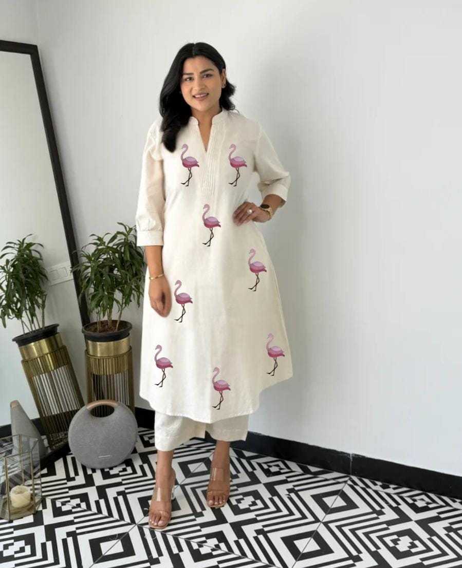Woman wearing a white dress with flamingo patterns in a room with plants and a mirror.