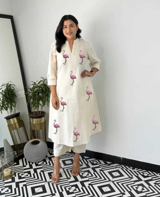 Woman wearing a white dress with flamingo patterns in a room with plants and a mirror.