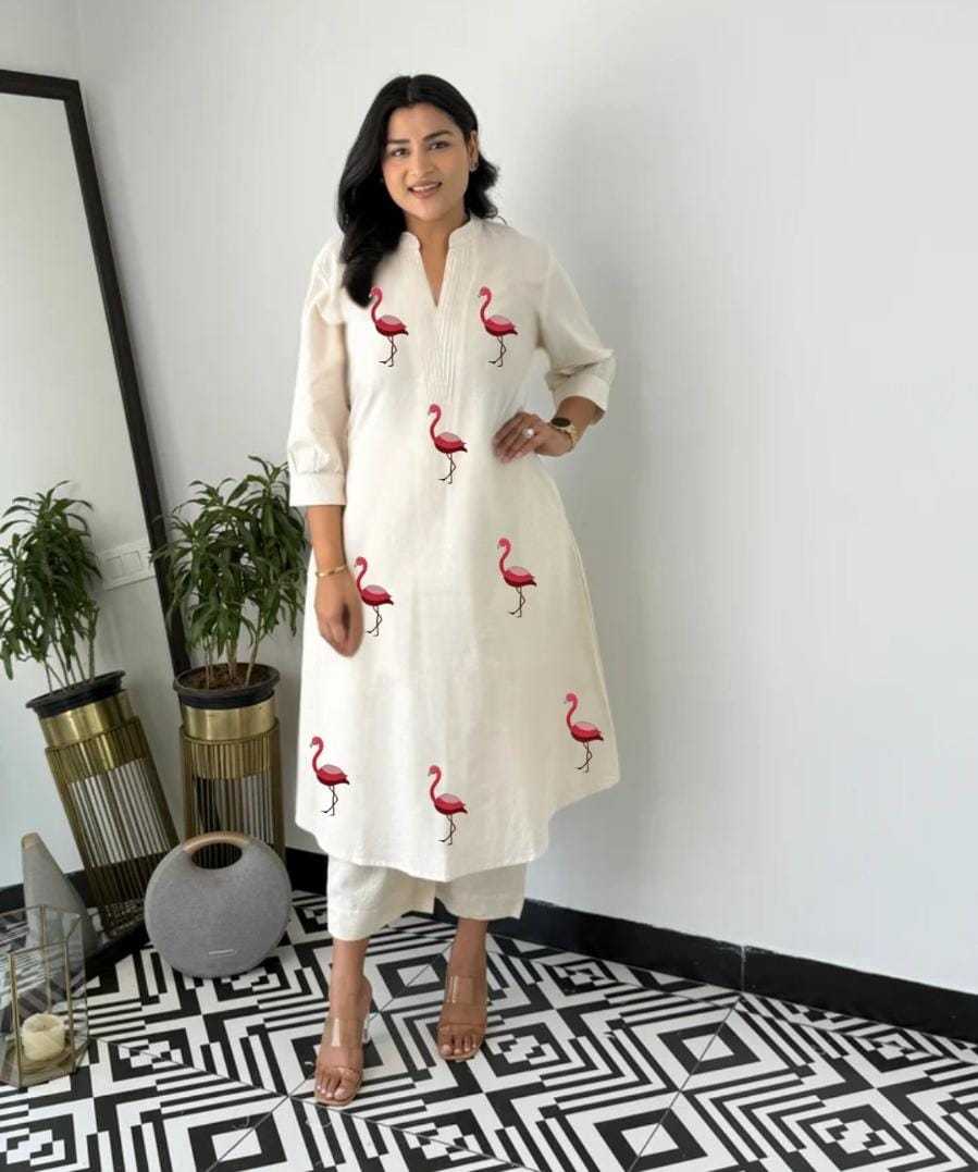 Woman wearing a white dress with pink flamingo patterns in a room with plants and decorative items.