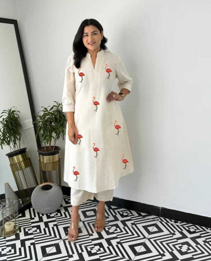 Woman wearing a white kurta with red flamingo patterns in a room with decorative plants and a mirror.