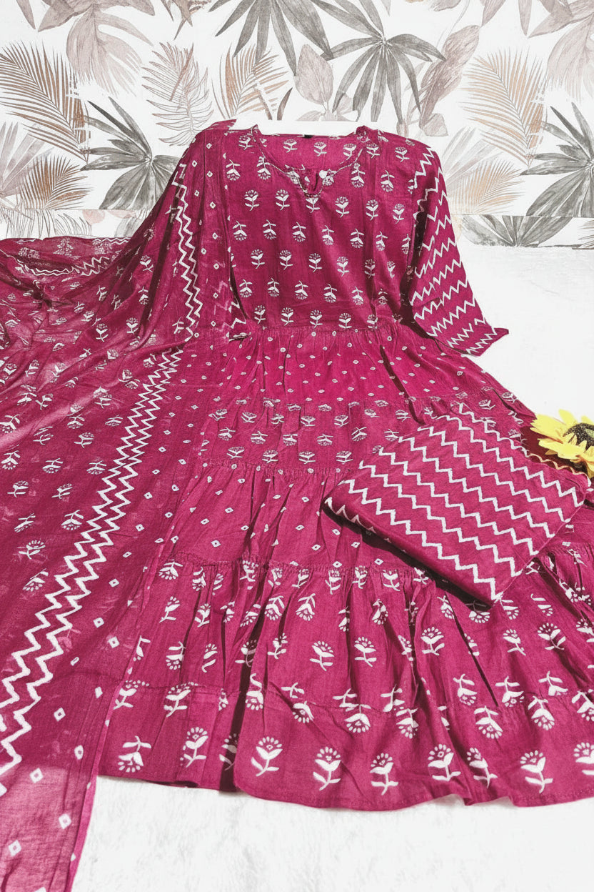 A magenta pink three-piece women's salwar suit set with white block prints, featuring a flared kurta, matching pants, and a dupatta with chevron borders, laid flat on a light surface with a tropical patterned backdrop.